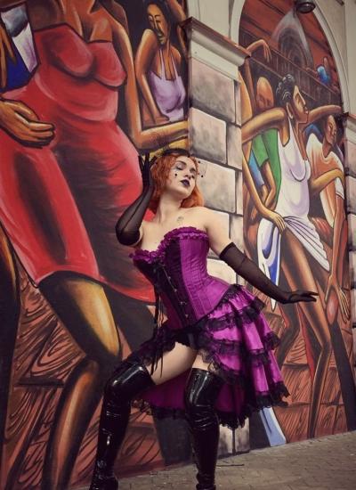Gothic Amethyst Burlesque Жіночий костюм кабаре: фіолетовий атласний корсет, ярусна спідниця з чорним мереживом і лакові ботфорти біля муралу