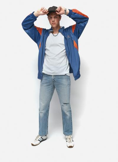 Tracksuit Street Legend У повний зріст, руки на козирку кашкета: розстебнута синя олімпійка з помаранчевими вставками, годинник, джинси з дірою на коліні