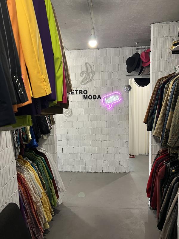 Біла цегляна стіна з вивіскою RETRO MODA і неоновим написом hello, по боках вішаки з піджаками і сорочками